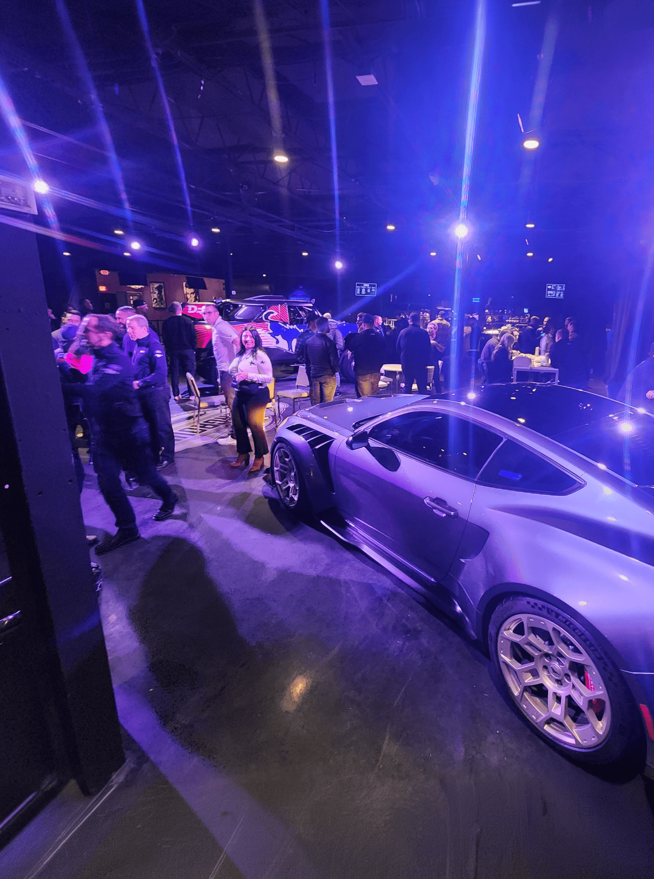 Purple-lit indoor event with a crowd mingling behind a silver wide-body coupe in the foreground