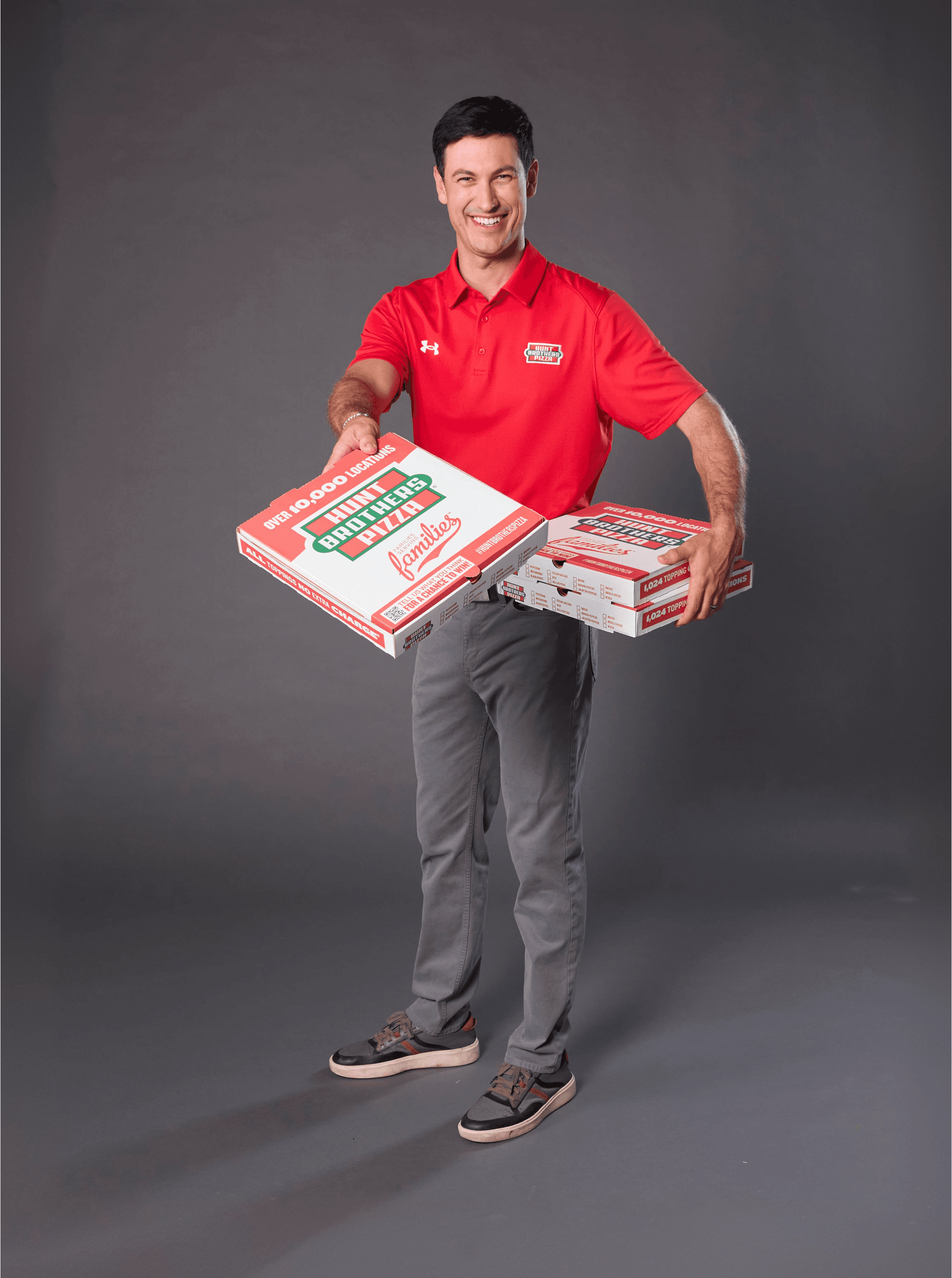 Man in a red polo handing over Hunt Brothers Pizza family sized boxes against a gray studio backdrop