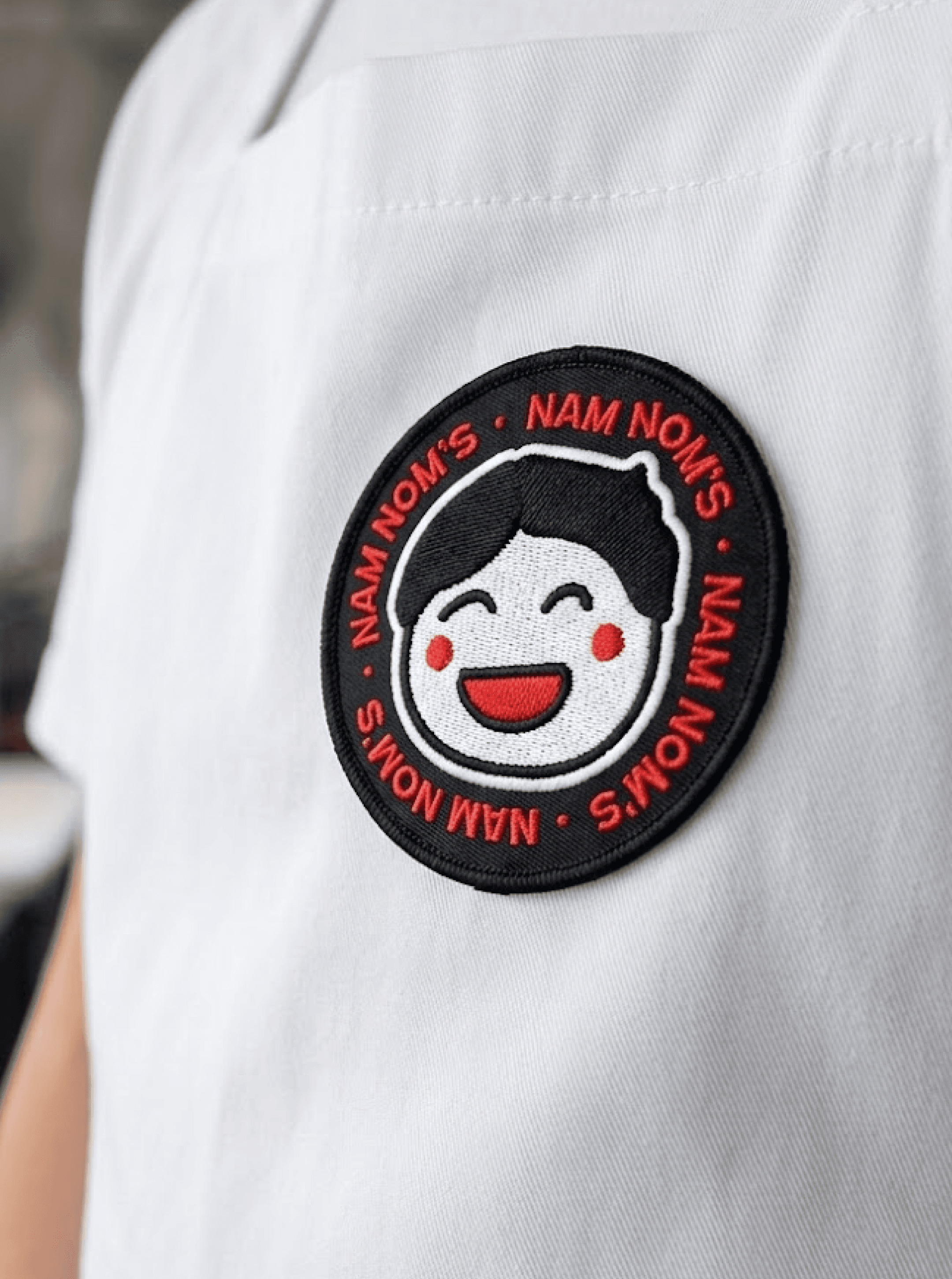 Embroidered patch on a white work shirt showing a smiling cartoon face ringed by the words "NAM NOM'S"