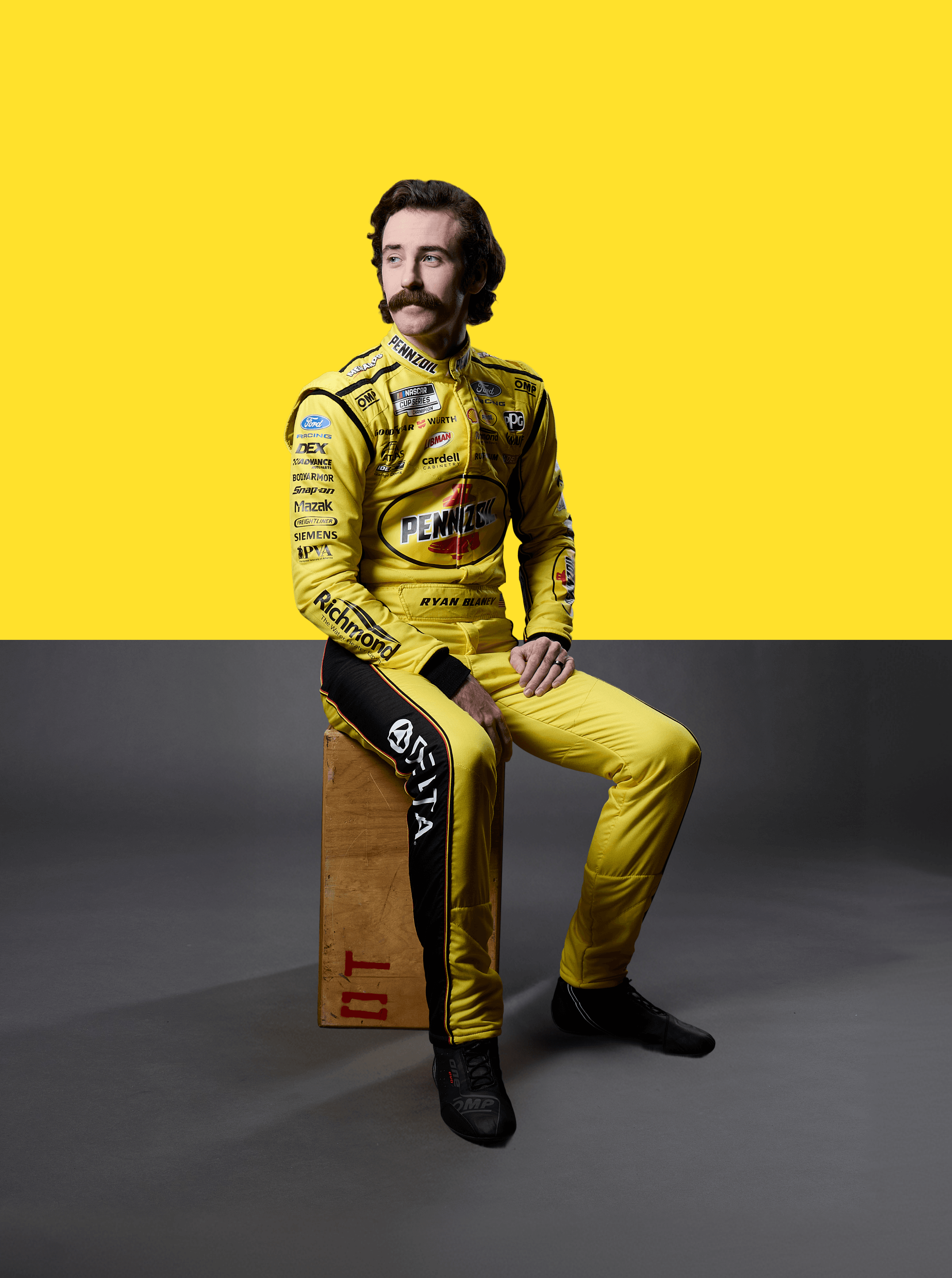 Mustached driver seated on a wooden crate in a yellow Pennzoil suit against a split yellow and grey wall