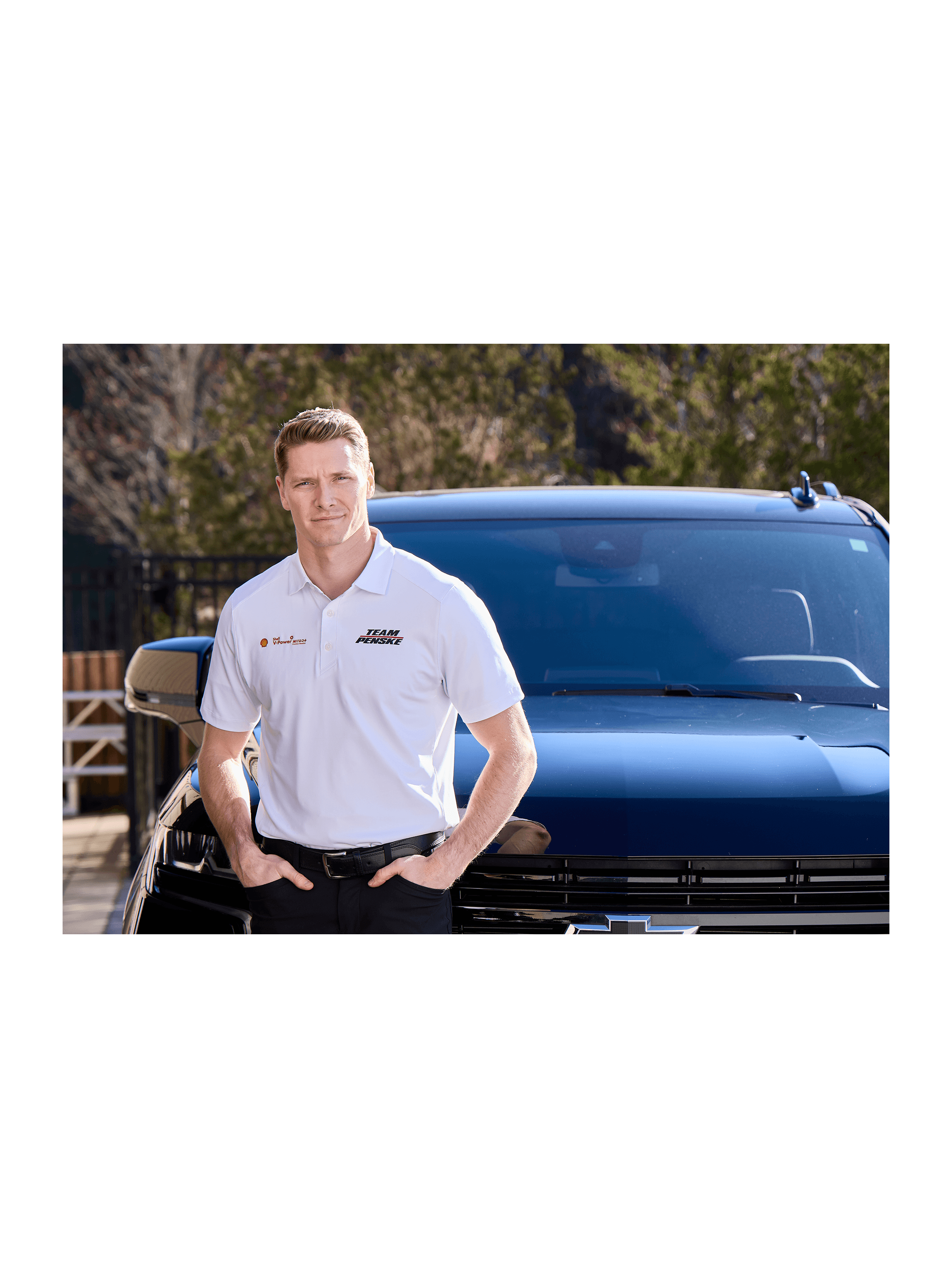 Man in a white Team Penske polo standing hands in pockets in front of a blue Chevrolet pickup outdoors