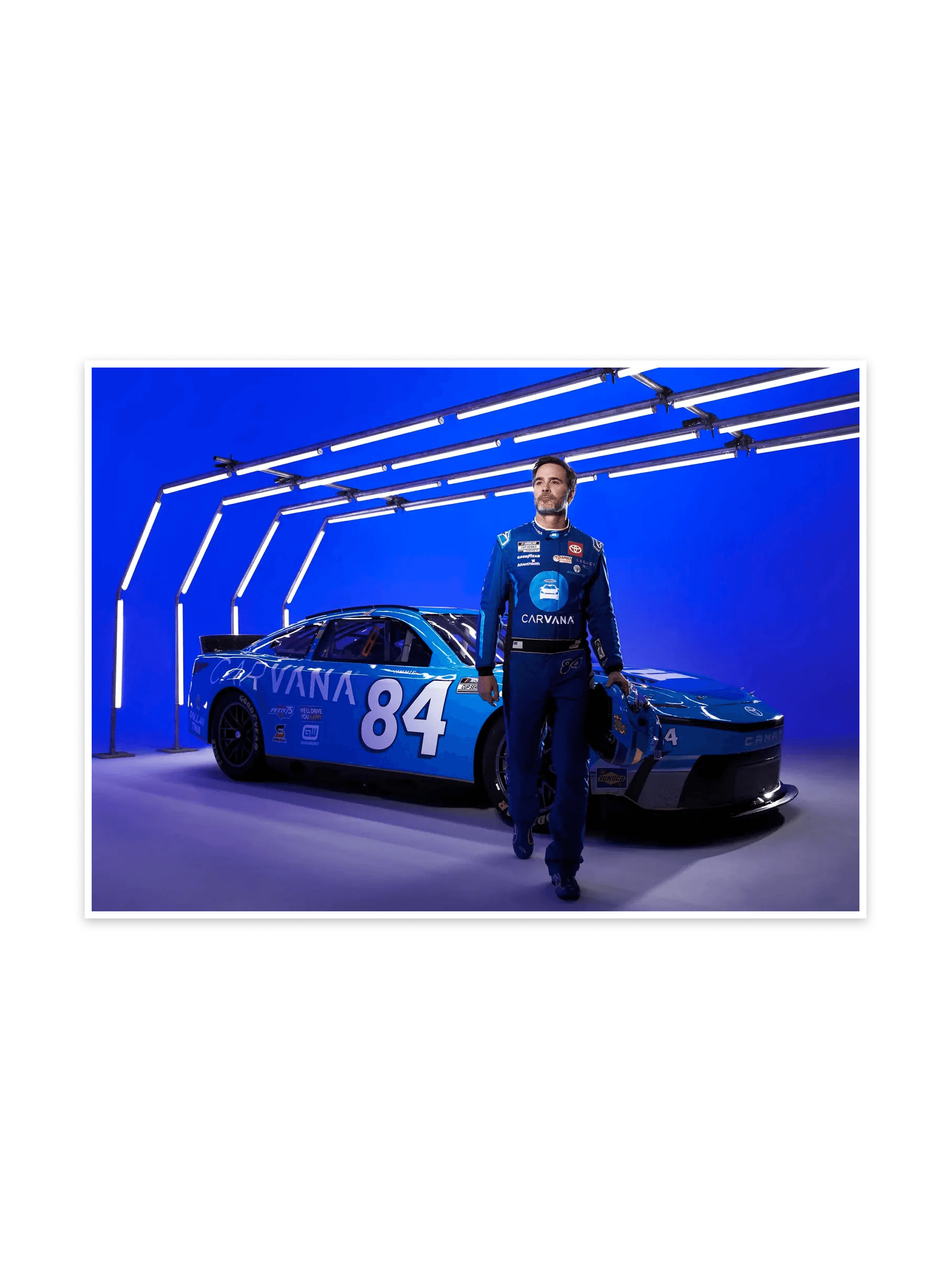 Driver in blue Carvana gear walking past the number 84 Carvana Cup car under white LED tubes