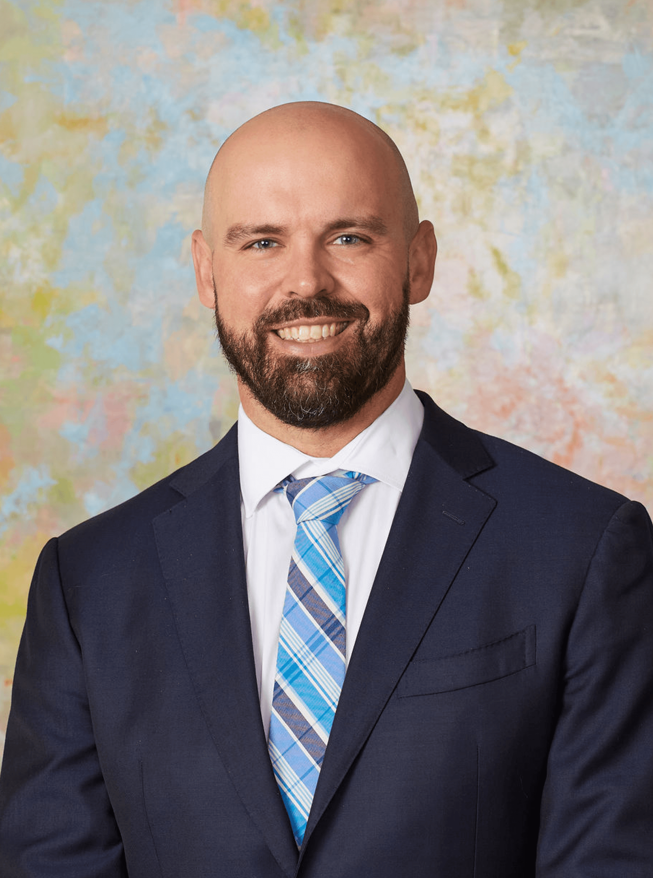 Bald man with a dark beard in a navy suit and blue plaid tie, smiling against a pastel backdrop