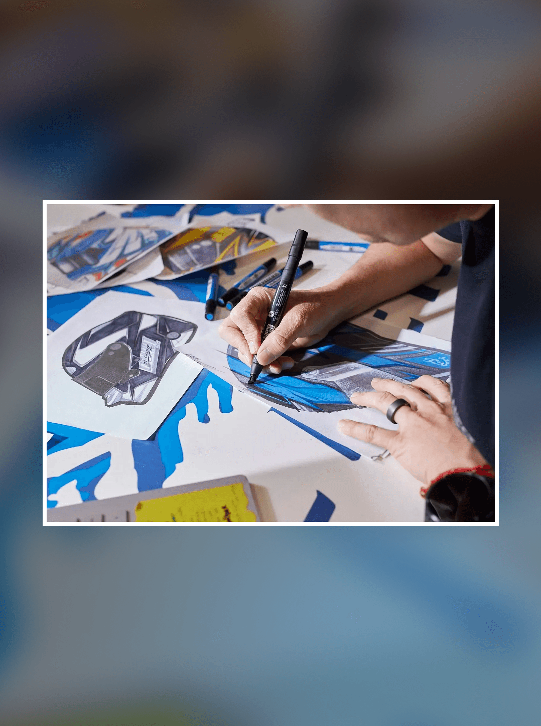 Hand with a black ring inking a blue and white helmet design in marker, more sketches spread around