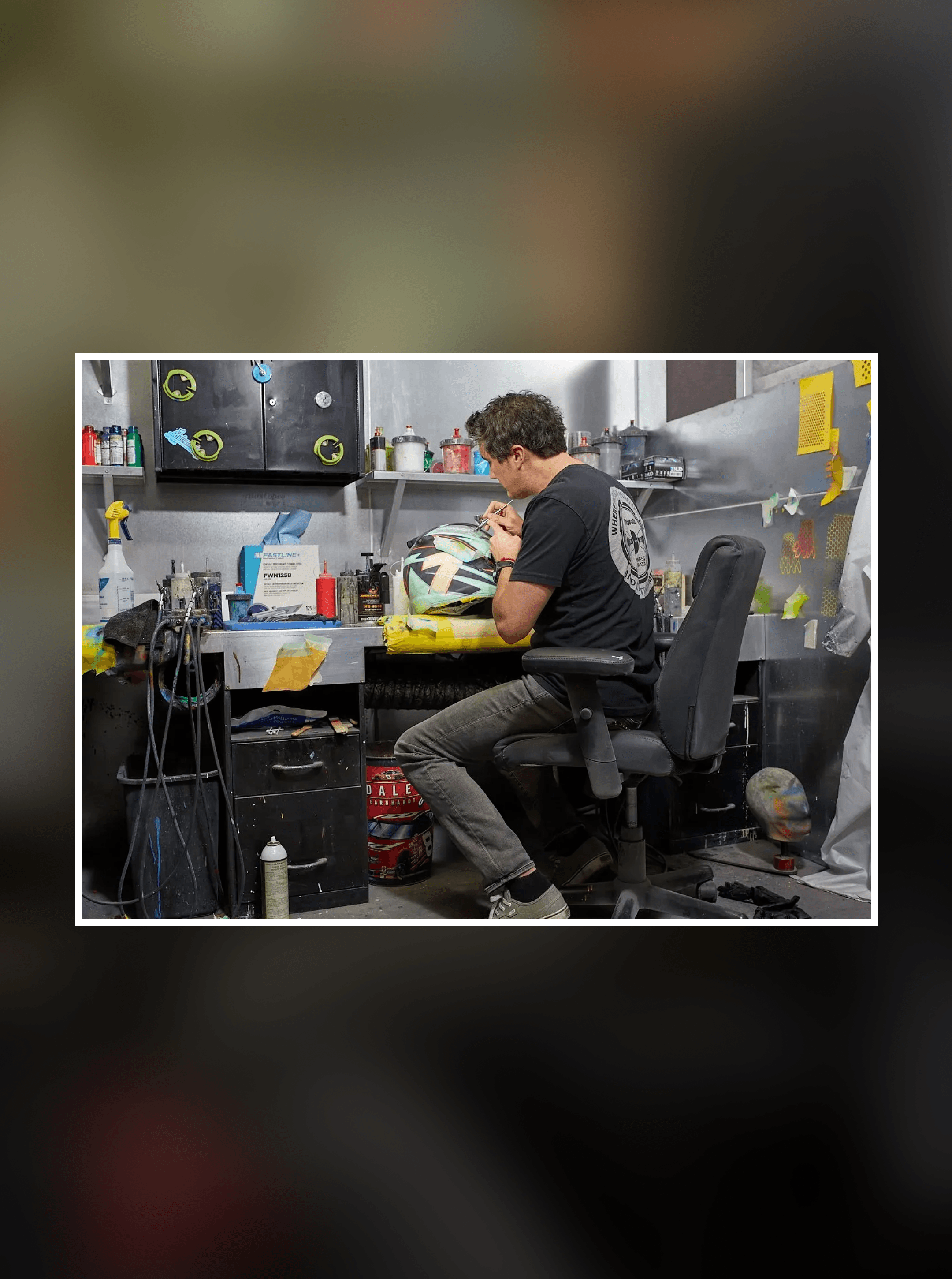Wide view of the cluttered paint shop with the artist seated at his bench holding a yellow and teal helmet
