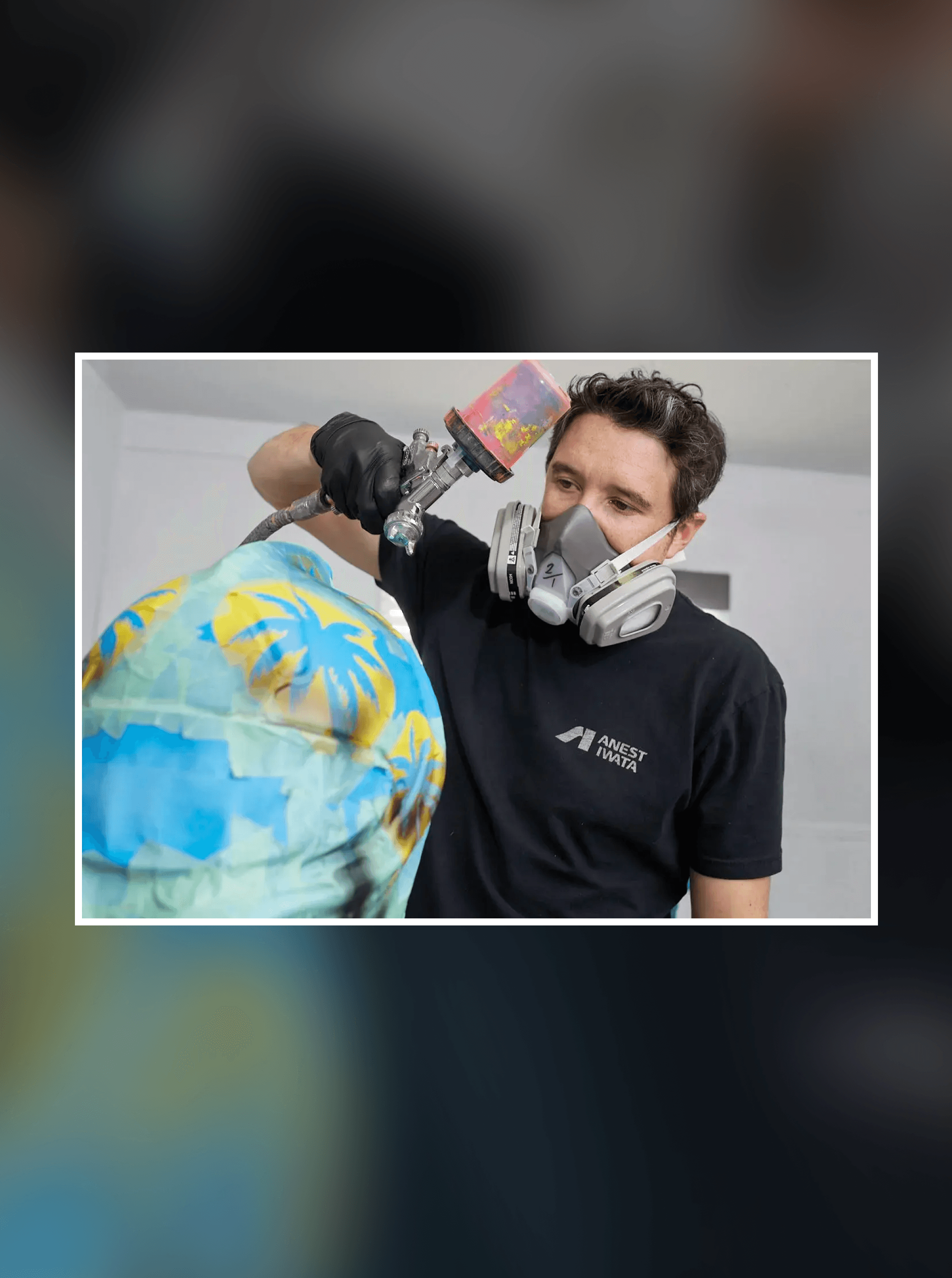 Painter in a respirator spraying a light blue helmet with yellow palm trees using a handheld spray gun