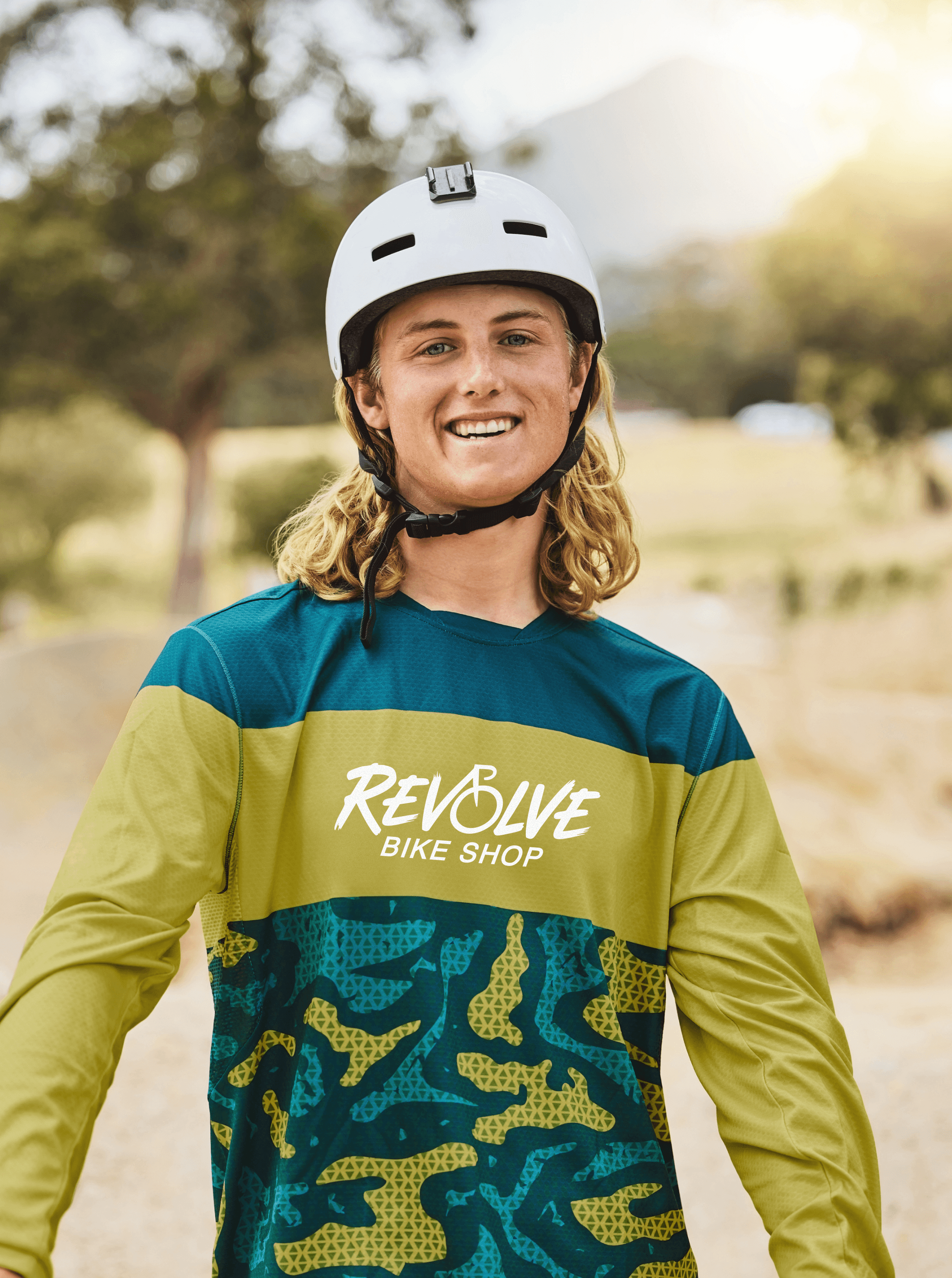 Young rider in a white helmet smiling in a teal and olive Revolve Bike Shop jersey with camo panel