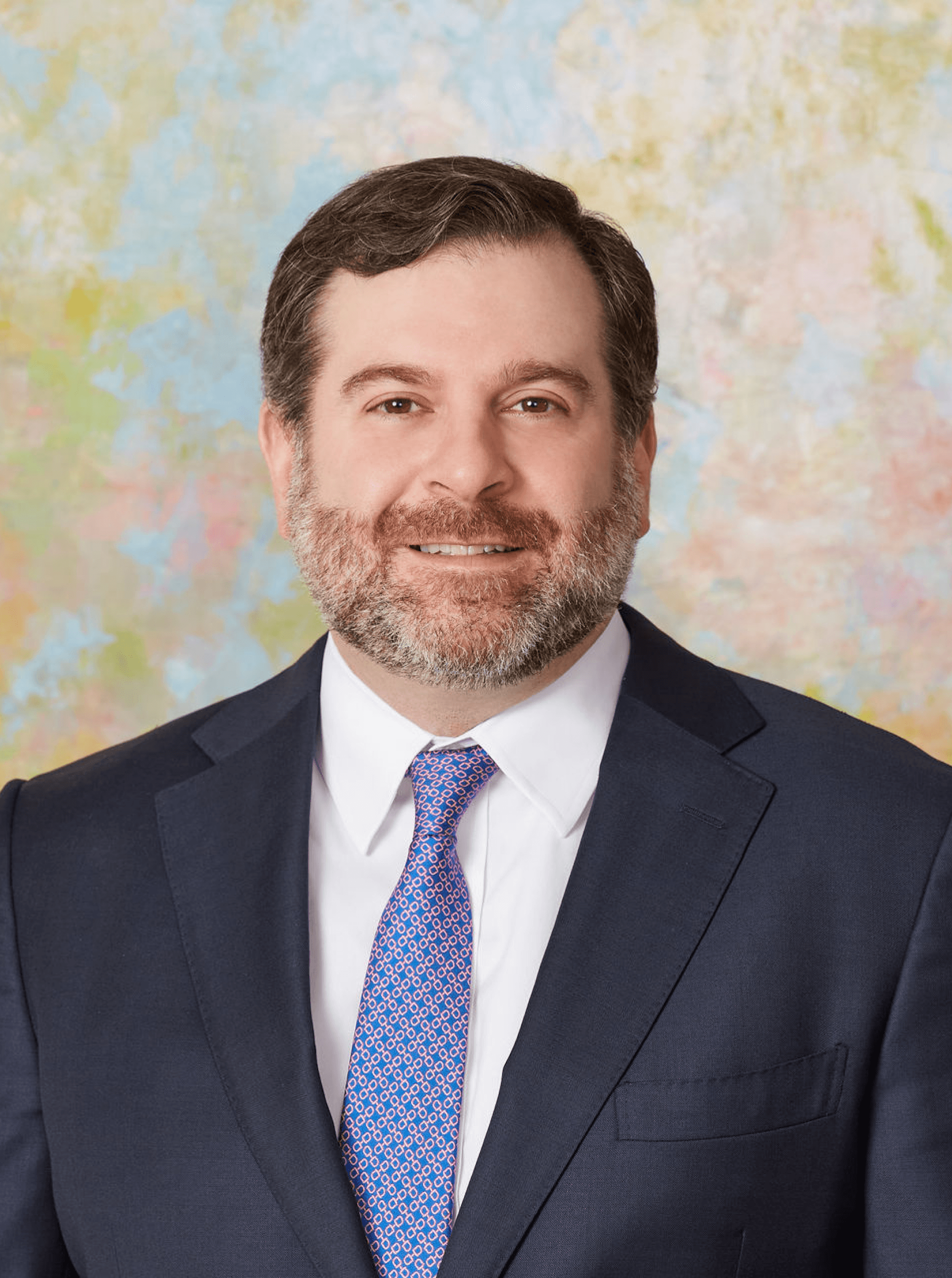Bearded man in a dark navy suit with a red and blue patterned tie, pastel studio backdrop behind