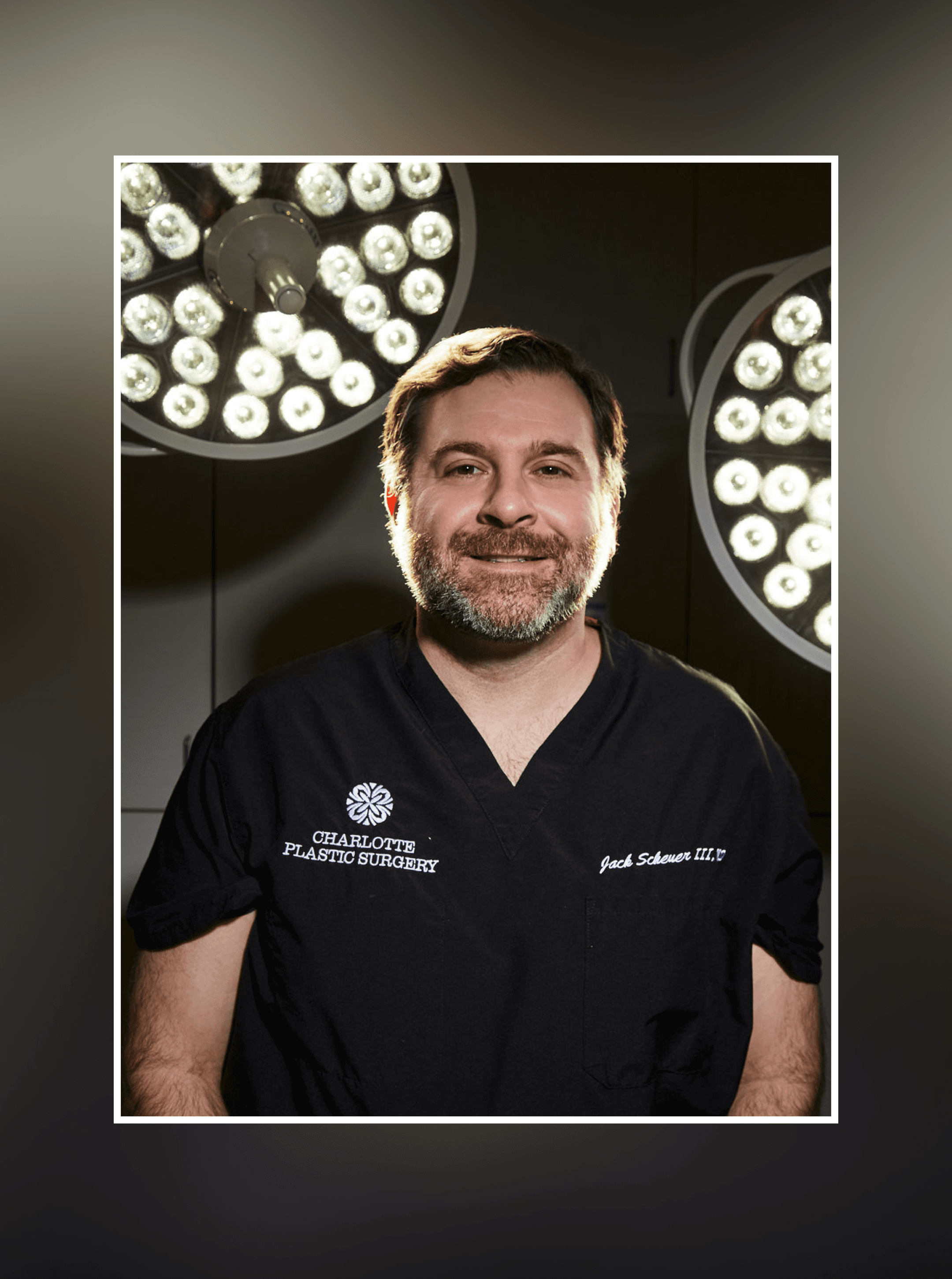 Bearded surgeon in black scrubs embroidered Charlotte Plastic Surgery, framed by two surgical light heads
