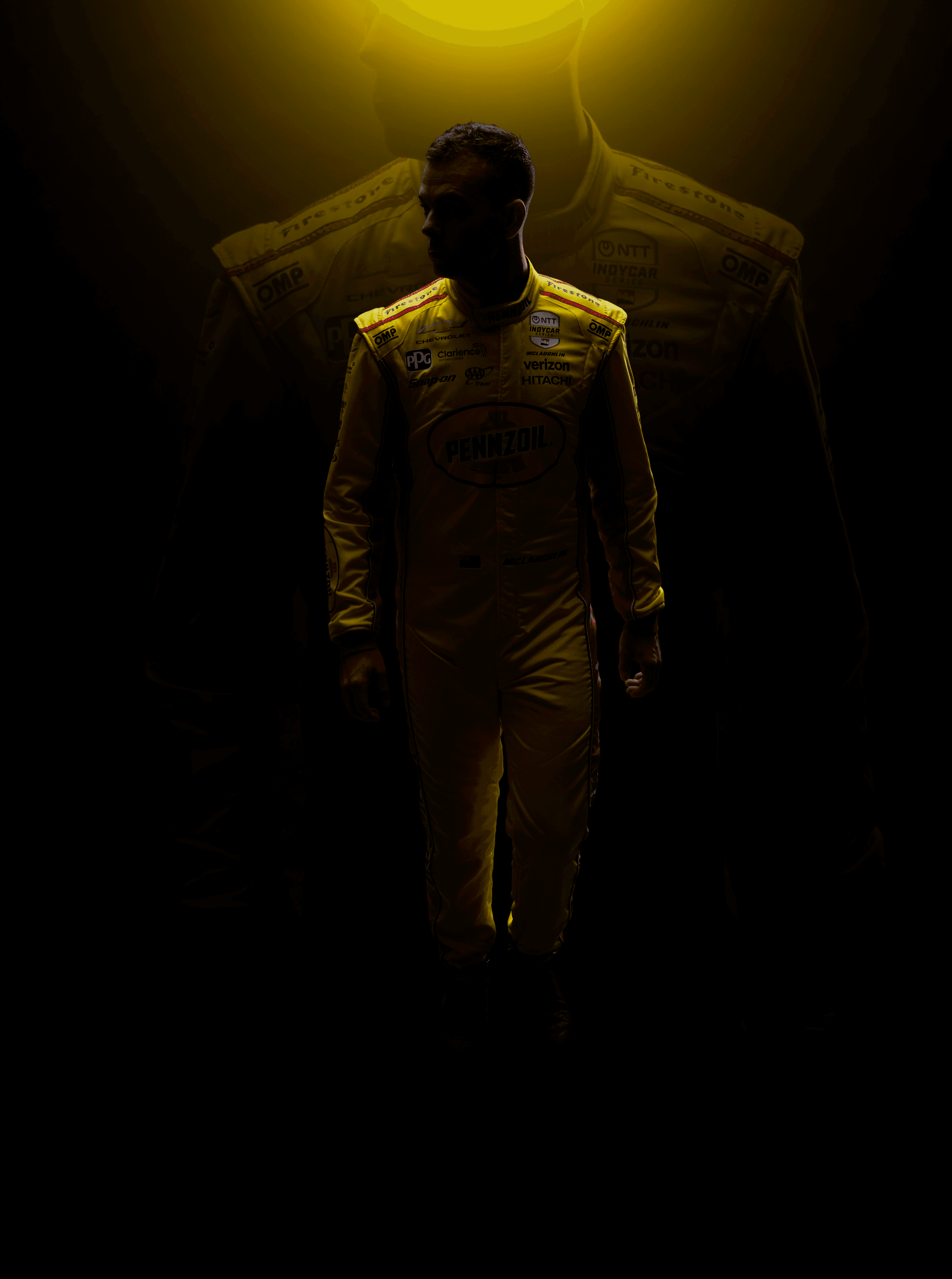 Driver in a yellow Pennzoil fire suit walking out of darkness with two hanging suits backlit behind