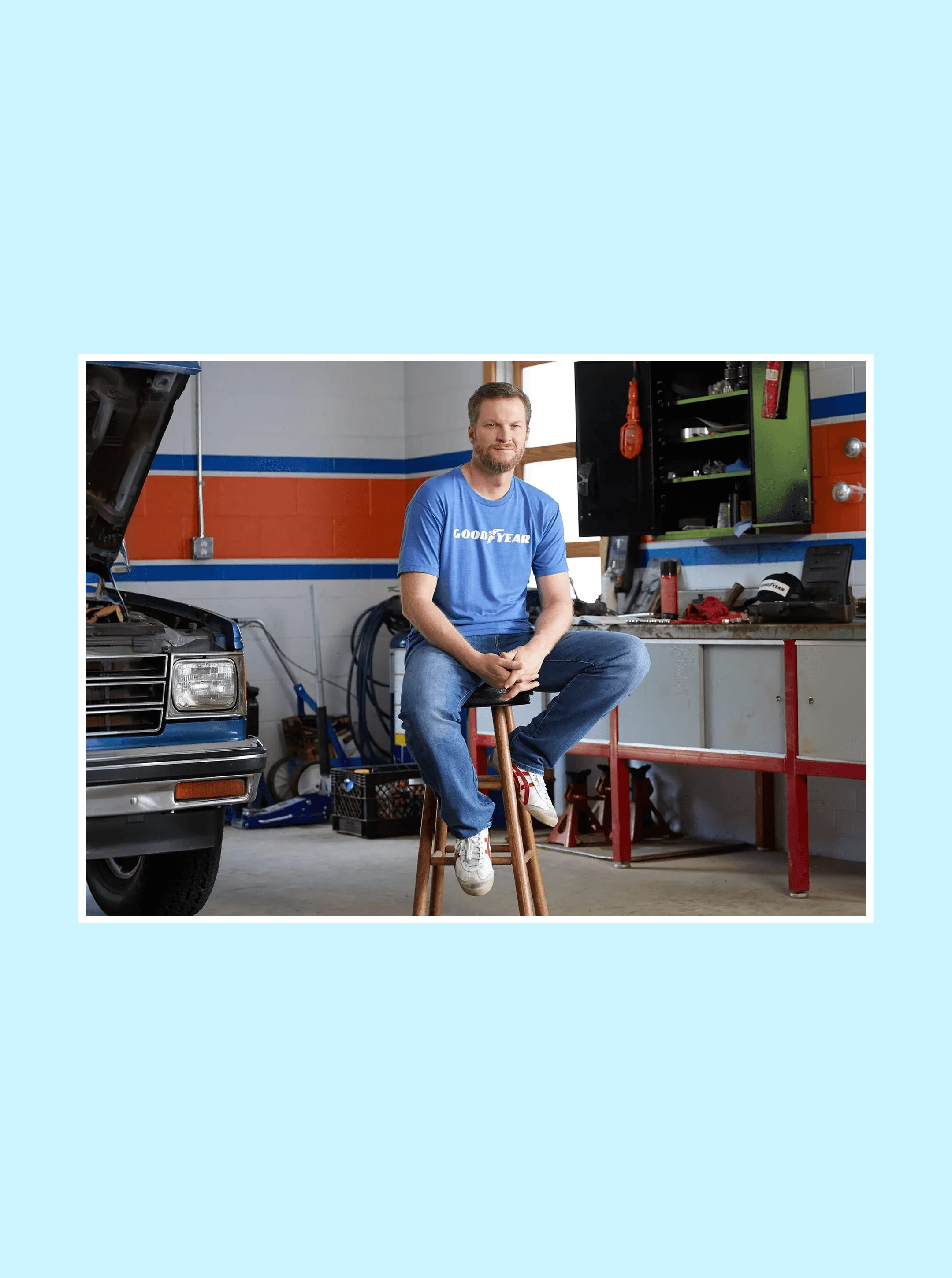 Man in a Goodyear T-shirt seated on a wooden stool in a garage beside a truck with its hood raised