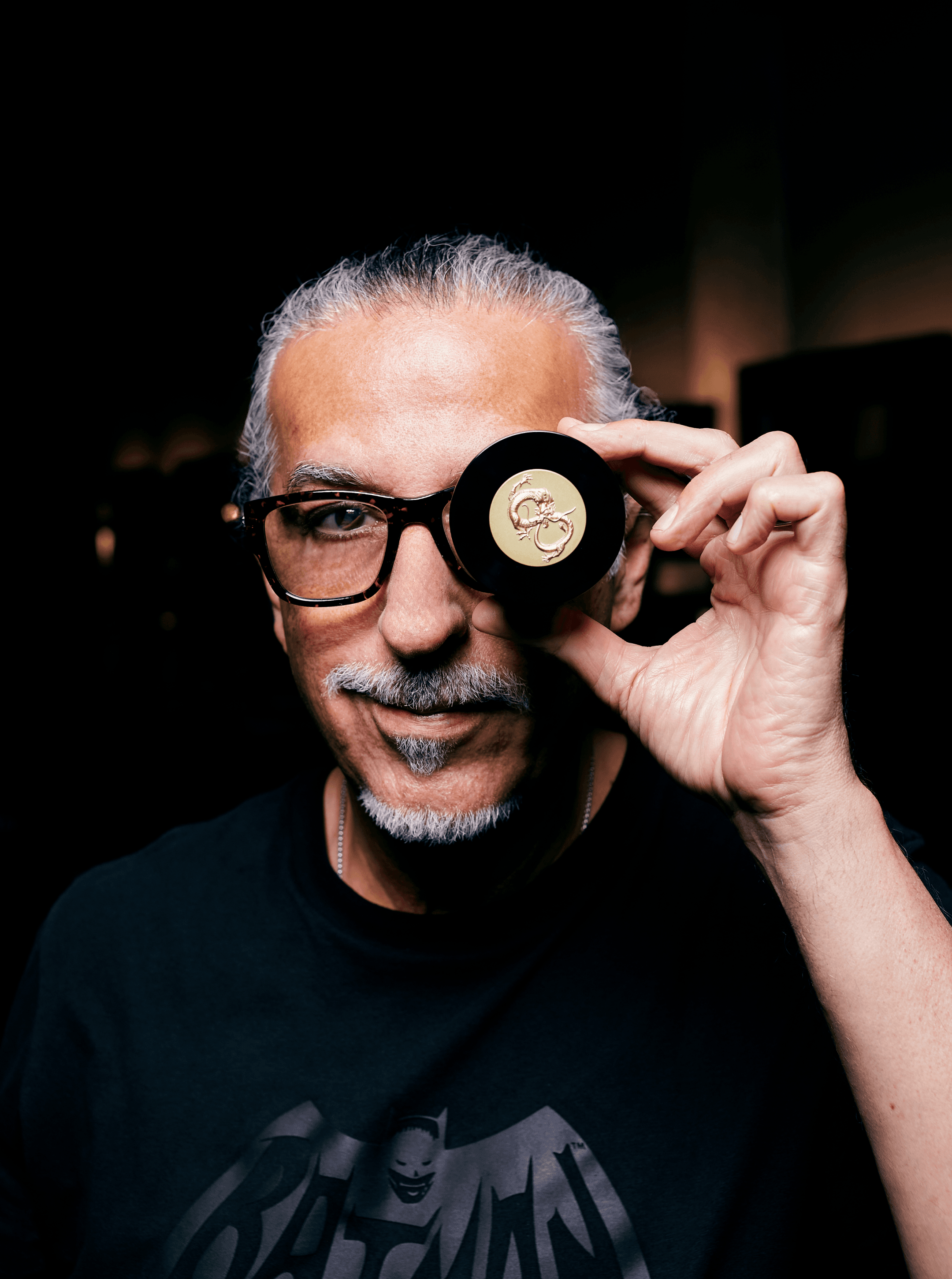 Gray-haired man in glasses and a Batman tee holding a black AZ cap with a gold logo over one eye