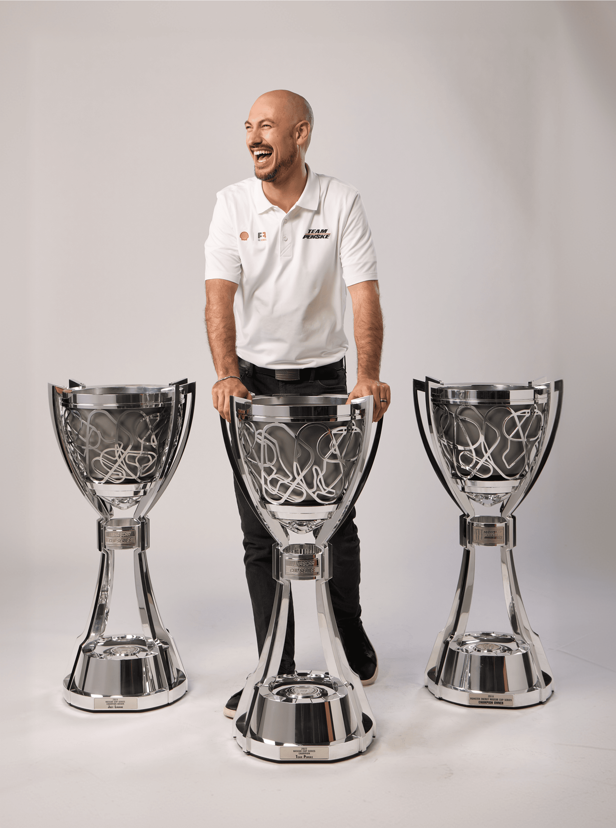 Man in a white Team Penske polo laughing behind three tall chrome championship trophies