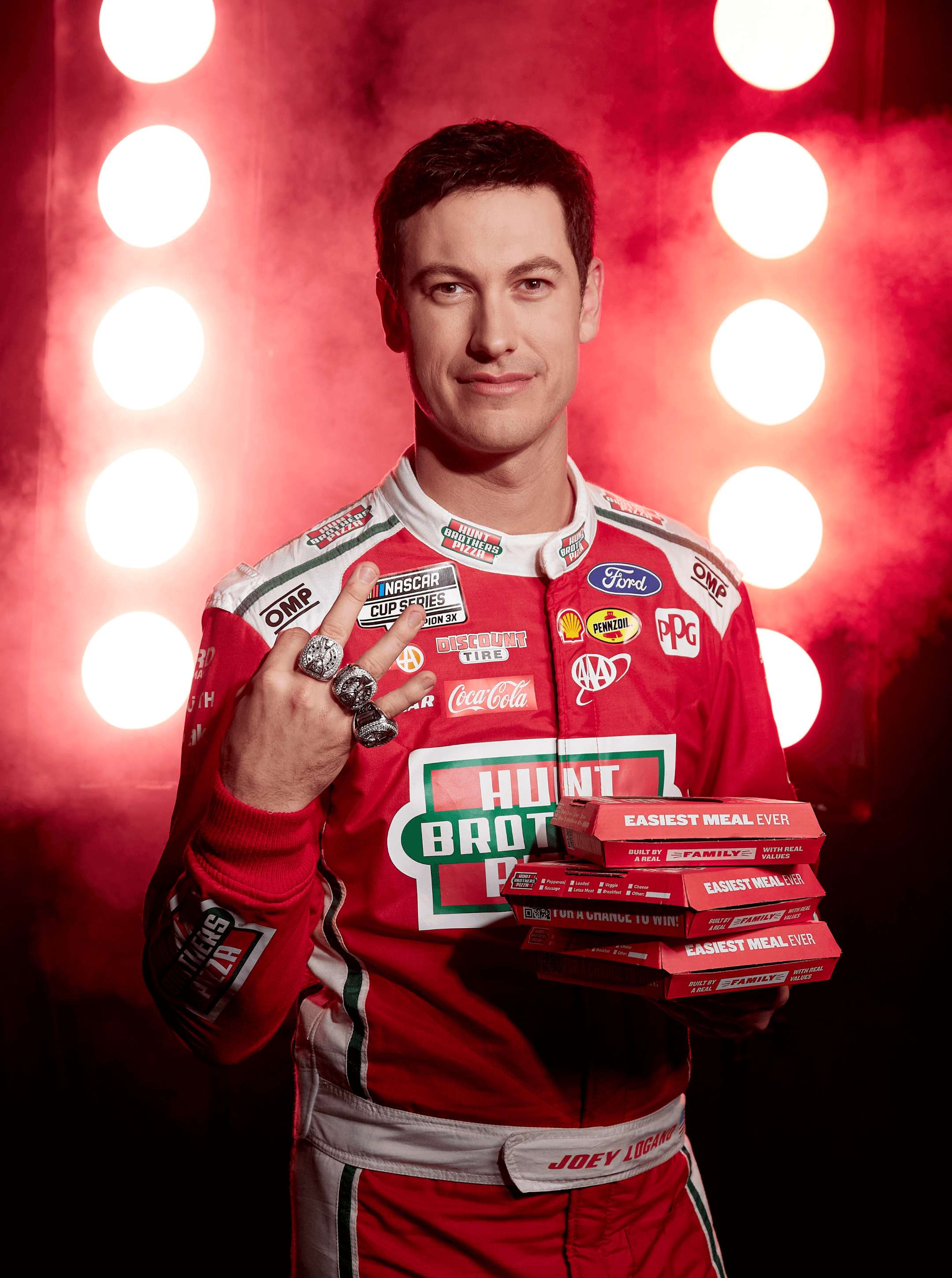 Joey Logano holding a stack of Hunt Brothers pizza boxes marked Easiest Meal Ever while showing his rings